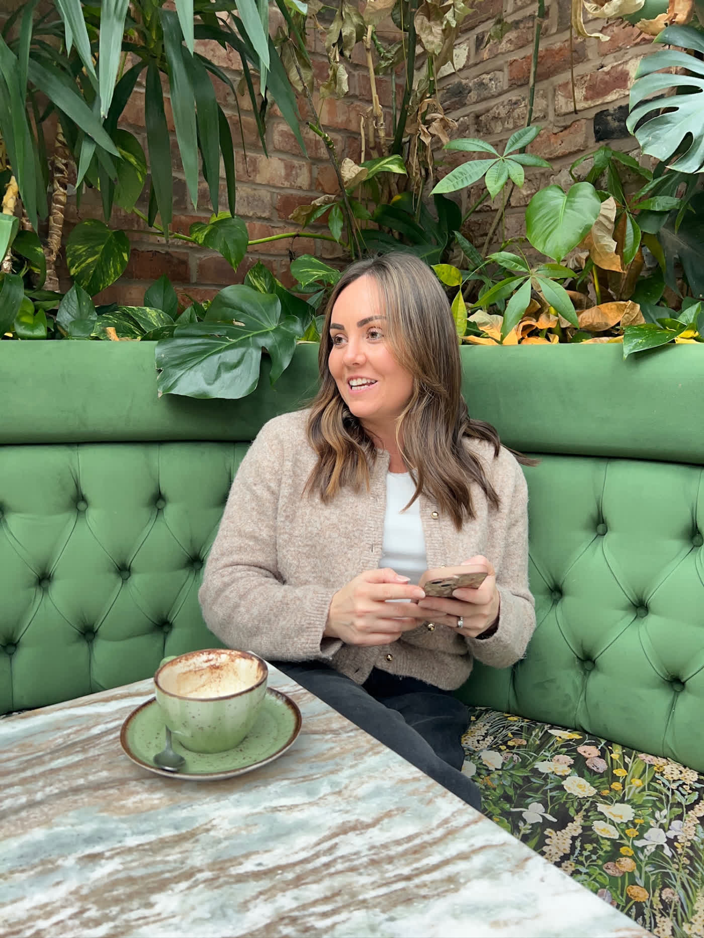 Leigh-Anne smiling in a café booth surrounded by plants, phone in hand